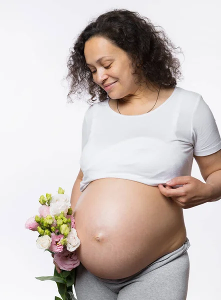 美丽的多民族快乐的胖女人 期待着的成年母亲抱着一束春花 在怀孕后期微笑着看着自己怀孕的肚子 在白色的工作室背景下与世隔绝 — 图库照片