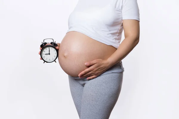美しい裸の腹を持つ妊婦の白い背景にクローズアップスタジオショット 目覚まし時計を保持 生物時計の音 出産の概念 出生の予定日 — ストック写真
