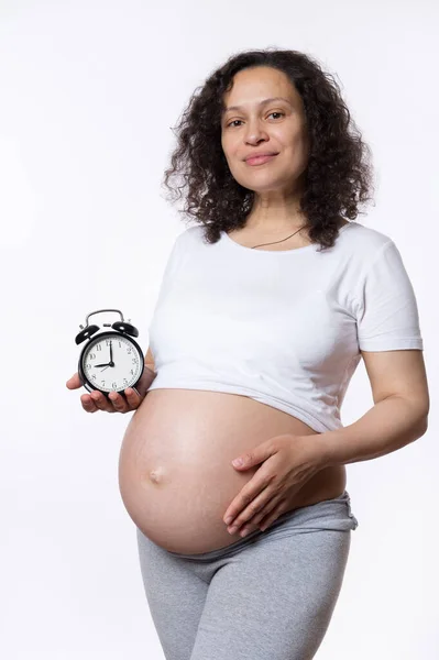 魅力的な妊婦は優しく彼女の腹をストローク かなり笑顔 子供の誕生の期待される日付に生物学的時間とカウントダウンをカチカチとカメラに目覚まし時計を示しています 赤ちゃんを待ってる — ストック写真