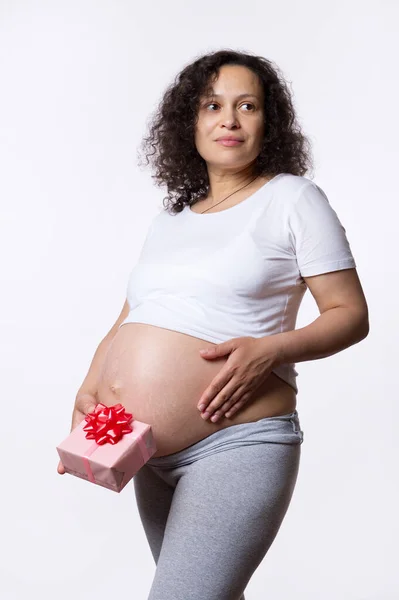 美丽的卷曲的多民族孕妇 准妈妈 心事重重地把目光投向一边 摸着肚子 把可爱的礼品盒放在她赤身裸体的腹部 与白色隔离 母亲节快乐的概念 — 图库照片