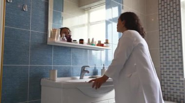 Çok etnik, orta yaşlı kıvırcık saçlı, beyaz waffle bornozlu hamile kadın banyo aynasında kendine hayran. Sabah rutinleri. Hamilelik zamanında kaplıca ve güzellik prosedürleri
