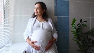 Enfes kıvırcık saçlı, kıvırcık saçlı, yetişkin, güzel bir kadın hamile karnını hafifçe okşuyor ve ev tuvaletinin penceresinde duruyor. Dertsiz mutlu hamilelik ve annelik kavramı