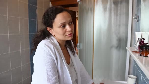 一个迷人的中年多种族女人的特写 她看着镜子里的倒影 触摸着自己的脸和皱纹 站在家里的包厢里 穿着白色的华夫饼浴衣 — 图库视频影像