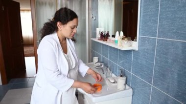 Gecelikli ve beyaz waffle bornozlu orta yaşlı hamile kadın, göz bantlarının altında hidrojenle bir kavanoz açıyor, ev banyosunun içinde duruyor. Hamilelik zamanı cilt bakımı kavramı