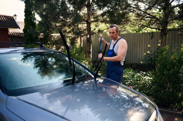 stock image Handsome Caucasian man, car service worker in blue working uniform, directs sprayer with pressurized water jet at windshield and wipers, washes a vehicle with modern high pressure cleaning equipment