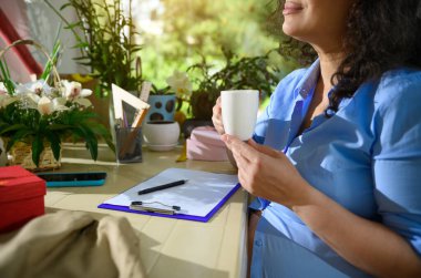 Yakın plan hamile kadın, etkinlik yöneticisi, elinde beyaz bir bardak sıcak içecek tutan çiçekçi, elinde dosya ve siparişlerle masasında oturuyor, çiçek düzenlemeleri ve çiçek tasarım stüdyosundaki hediyelerle çevrili.