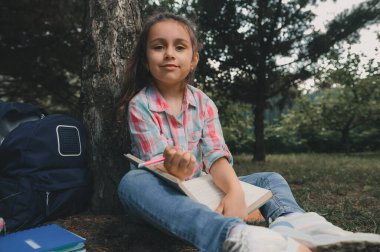 Beyaz, sevimli küçük kız çocuğu, pembe kareli gömlek ve kot pantolon giyen okul çocuğu, parktaki ağacın altında oturuyor, elinde bir ders kitabı ve kalem, gülümsüyor, kameraya güvenle bakıyor.