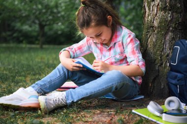 Beyaz, tapılası birinci sınıf öğrencisi, şık pembe ve turkuaz ekose gömlek ve mavi kot giyen ilkokul öğrencisi, şehir parkındaki bir ağacın altında rahatça oturup ödev yapıyor..