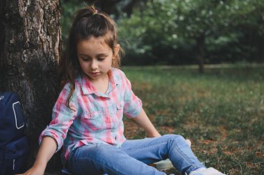 At kuyruklu, ekose gömlekli ve mavi kotlu şirin bir kız parkta dinleniyor, yeşil çimenlerde oturuyor, ağaca yaslanıyor ve okuldan sonra oynuyor. - İnsanlar. Çocukluk. Çocuklar işte. Yaşam biçimi