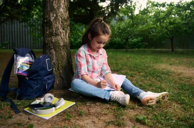 Küçük sevimli beyaz kız parkta ödev yapıyor, bir deftere yazı yazıyor, okul malzemeleriyle dolu bir sırt çantasıyla bir ağacın altında oturuyor. Eğitim. Öğreniyorum. Mutlu çocuklar. Çocukluk