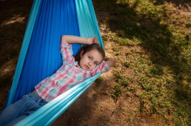 Kafkasyalı küçük sevimli kız çocuğu dışarıda dinleniyor, kameraya bakıyor, güneşli bir günde hamakta uzanıyor, yaz kampında ya da ormanda dinleniyor. Çocukluk. Eğlence için. Tatillerde. Tatiller.