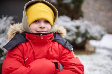 Beyaz tenli, yakışıklı, 11-13 yaşlarında, sıcak kırmızı ceketli ve sarı yün şapkalı bir gencin yakın plan portresi. Kameraya bakıyor, karla kaplı arka bahçede duruyor. Çocuklar işte. Çocukluk