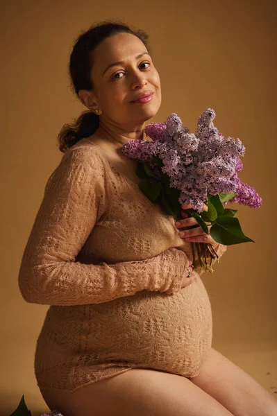 穿着漂亮的米色针织毛衣的漂亮的怀孕的快乐女人 在镜头前甜美地微笑着 摆出一束芬芳芬芳的百合花 坐在一个米色的孤零零的工作室背景上 — 图库照片