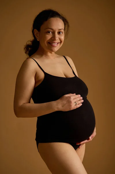 多民族のブルネット 黒い下着の幸せな灰色の女性は 彼女の大きな妊娠中の腹を愛撫し カメラを見て笑って ベージュのスタジオの背景に孤立した 妊娠中のファッション 産前産後ケア — ストック写真