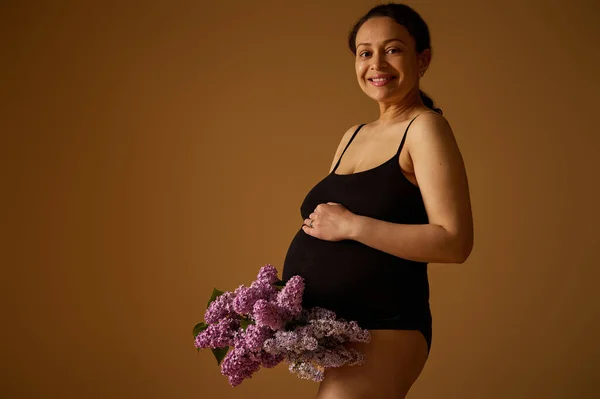 多民族妊娠中の女性のスタジオの肖像画腹を保持し カメラを見て ベージュの背景にライラックを咲かせてポーズ 体のポジティビティ 皮膚の不完全さ ストレッチマーク — ストック写真