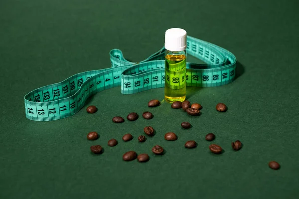Still life with a transparent bottle of massage anti-cellulite oil, measure tape and coffee beans scattered on isolated green background. Slimming and weight loss concept. Cosmetology. Copy ad space