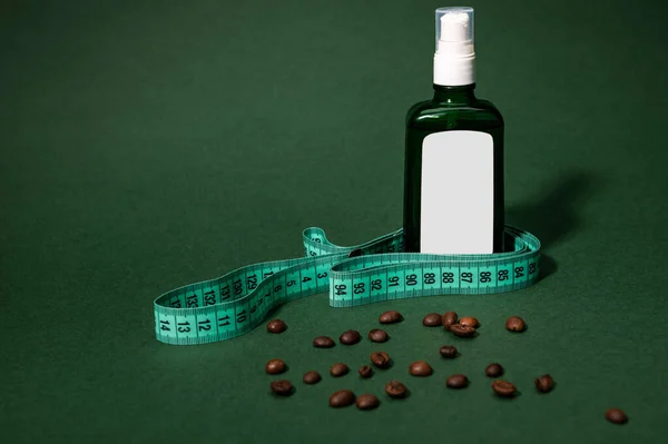 Still life. Cosmetic bottle from dark glass with a massage oil, measure tape and coffee beans scattered on green background. Anti-cellulite, weight loss, blood circulation improvement and spa concept.