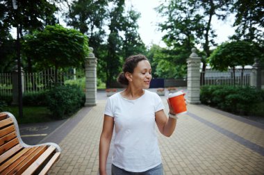 Çevresel kağıt bardakta kahve içen, gülümseyen, yaz günü şehir parkında yürürken yan yana bakan göz kamaştırıcı Latin Amerikalı genç kadın. - İnsanlar. Yaşam tarzı. Boş zaman aktivitesi