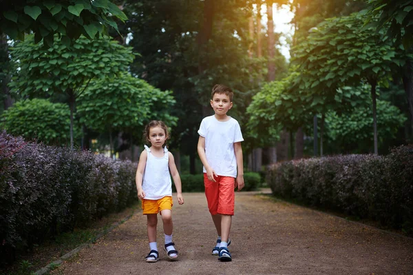 stock image Adorable two kids, boy and girl, brother and sister in white t-shirt and orange shorts, walking along the alley of a park at sunset on a sunny summer day. Childhood. Children. Recreation. Lifestyle