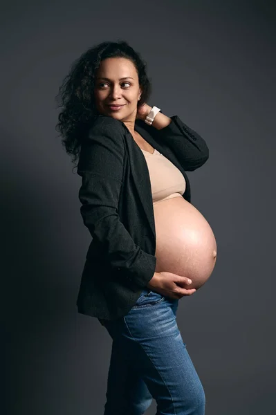 ぼかしアート 豪華なラテンアメリカの灰色の女性の本物の肖像画 優しく彼女の妊娠中の腹に触れ 脇を見て笑って エレガントなブレザーで身を包んだ 孤立したファッショングレーのスタジオの背景 — ストック写真