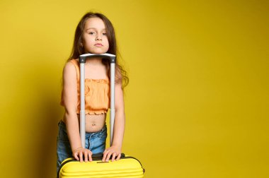 Beautiful Caucasian little girl in stylish bright summer orange top and casual blue jeans, standing near her vibrant trendy yellow suitcase, looking at camera, isolated on yellow studio background clipart