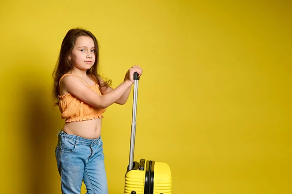長い髪の美しい小さな女の子は 青いジーンズとオレンジのトップに身を包んだ スーツケースを着て カメラを見て 黄色のスタジオの背景に隔離されたポーズ 旅行だ 旅行だ 週末旅行 — ストック写真