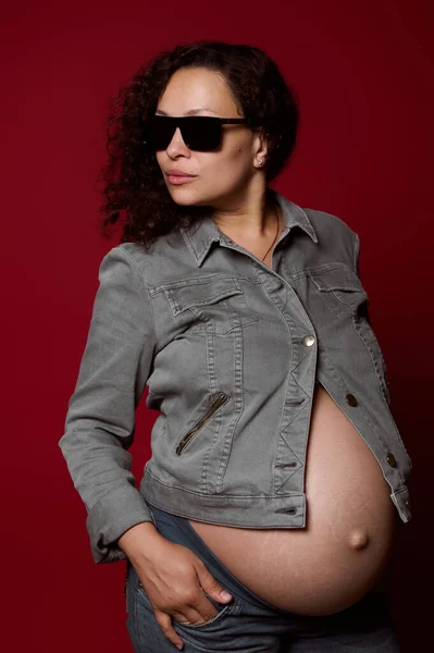 魅力的な巻き毛多民族の若い妊婦の広告スタジオショット30歳 流行のサングラスとカジュアルなデニムの服を着て 赤い色の背景に隔離されたポーズ — ストック写真