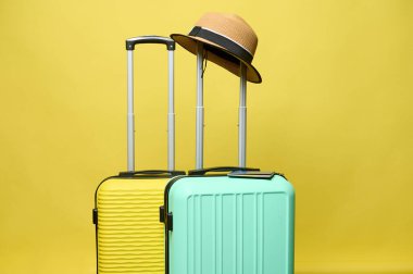 A summer straw hat on the handle of a light green suitcase, isolated over yellow studio background. Luggage. Low cost. Air flight. Travel. Tourism. Trip. Weekend getaway. Summer vacations. Holidays