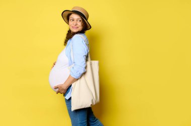 Authentic beautiful curly pregnant woman in straw hat and casual wear, holds her belly, smiles cutely looking aside at copy space, carrying eco linen shopping bag, isolated on yellow studio background
