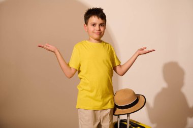 Beyaz tenli yakışıklı çocuk, sarı tişörtlü gezgin turist avuçlarında reklamlar için bir yer tutuyor, kameraya gülümsüyor beyaz stüdyo arka planında izole edilmiş bavulla ayakta duruyor.