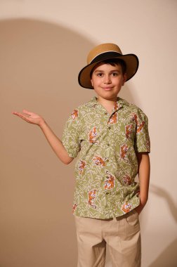 Hasır şapkalı, yaz desenli gömlekli, gri pantolonlu, hayali fotokopi alanını elinde tutan, kameraya kibarca gülümseyen, stüdyo arka planında izole edilmiş İspanyol turist çocuk.