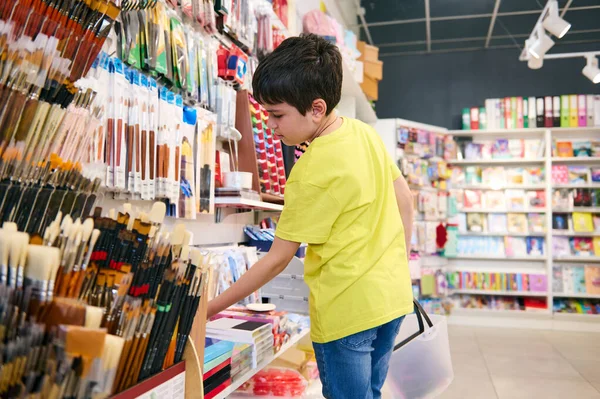 Latin American handsome teenage boy, primary school student, in blue denim and yellow t-shirt, holding shopping cart, standing in school stationery store, choosing school supplies. Back to school