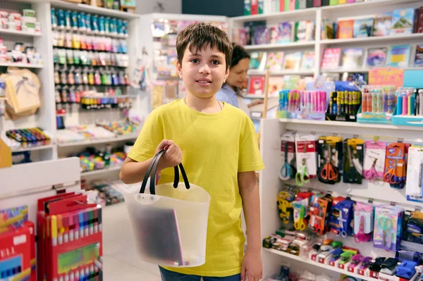 Handsome teen schoolboy smiles looking at camera, standing with a shopping cart in the school stationery store. Smart student boy buying school supplies. Back to school on new academic year semester