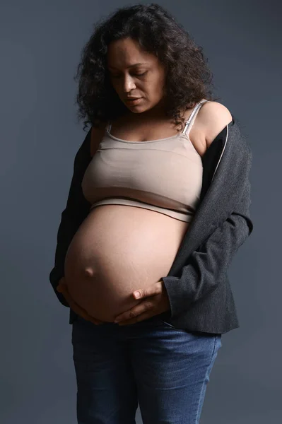穿着夹克和牛仔裤的漂亮女人 在她快乐无忧无虑的怀孕的第九个月里摸着怀孕的肚子 感受着孩子的脚踢 享受着产假 在灰色的工作室背景下与世隔绝 — 图库照片