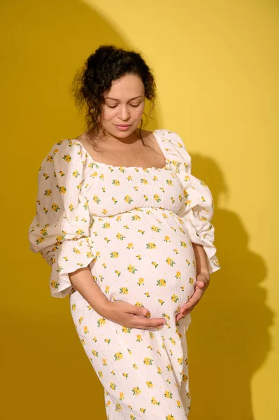 stock image Multi-ethnic curly haired middle aged pregnant woman, gravid expectant mother in stylish patterned summer sundress, touching her big belly, isolated over yellow studio background. Beautiful pregnancy