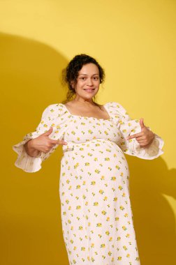 Hamile genç bir kadın 38 hafta sonra hamileliğinin sonlarına doğru koca göbeğini işaret ediyor. Mutlu, pozitif duygular yaşıyor, bebek bekliyor, kameraya bakıp gülümsüyor, sarı arka planda izole edilmiş..