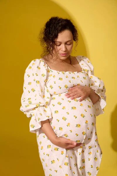 stock image Multiethnic curly haired middle aged beautiful pregnant woman putting hands on her belly, enjoying baby kicks, isolated over yellow background. Pregnancy and maternity. Human fertility. Women's health