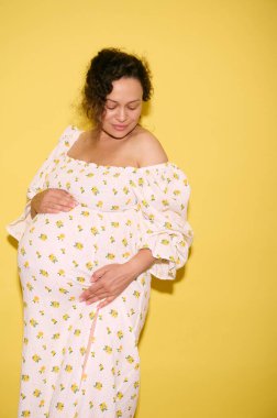 Çoklu etnik beden pozitif kıvırcık esmer, çekici, hamile bir kadın bebek bekliyor, koca göbeğini tutuyor, sarı stüdyo arka planında izole edilmiş.