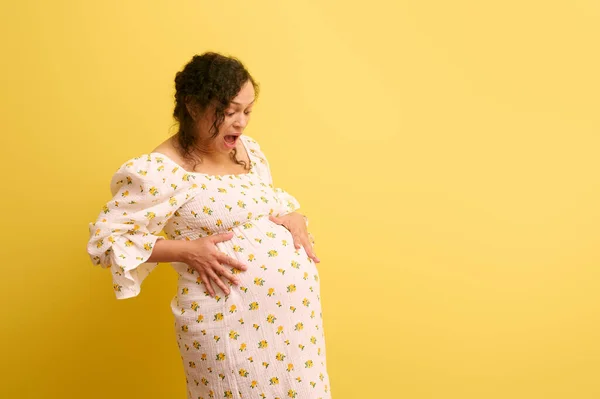 Curly ethnic pregnant woman, gravid female, mother to-be holding belly, crying and shouting, isolated on yellow background. Easy delivery, pregnancy and parturition concept. Obstetrics and gynecology