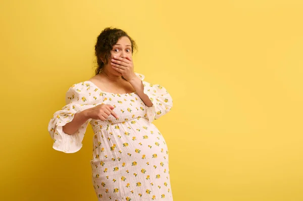 Easy delivery, pregnancy and parturition concept. Obstetrics and gynecology. Pregnant woman expressing shock, pointing at her belly, isolated over yellow studio background. Human fertility and health