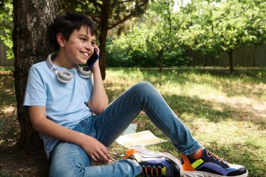 Neşeli yakışıklı genç çocuk cep telefonuyla konuşuyor, kulaklık takıyor, mavi tişört giyiyor ve şık bir kot pantolon giyiyor, parkta bir ağacın altında dinleniyor. Tonlanmış fotoğrafçılık. Çocuklar işte. Yaşam tarzı. Teknoloji