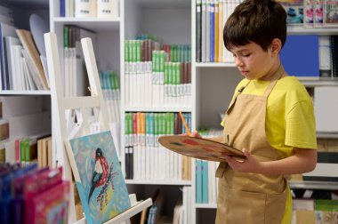 Önlüklü yakışıklı bir genç, boya fırçası ve palet kullanıyor, resim çiziyor, bir sanat atölyesinde ahşap sehpanın önünde duruyor, satılık tuvallerin arka planına karşı. Resim yapmak. Hobi olarak. Çocuklar.