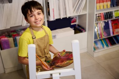 Yakışıklı ilkokul çocuğu yağlı boyalı bir palet tutuyor, resim sehpasında oturuyor, tuvale resim çiziyor, gülümsüyor, kameraya bakıyor, yaratıcı hobisinden hoşlanıyor, sanat stüdyosunda ilham arıyor.