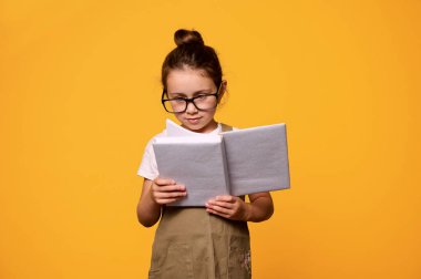 Gözlüklü zeki bir ilkokul öğrencisi, kitap okuyor, izole edilmiş turuncu bir geçmişi var. - İnsanlar. Yıkım. Dünya Kitapları Günü konsepti. Öğretmenler Günü. Akademik yılın yeni döneminde okula dönüyorum.