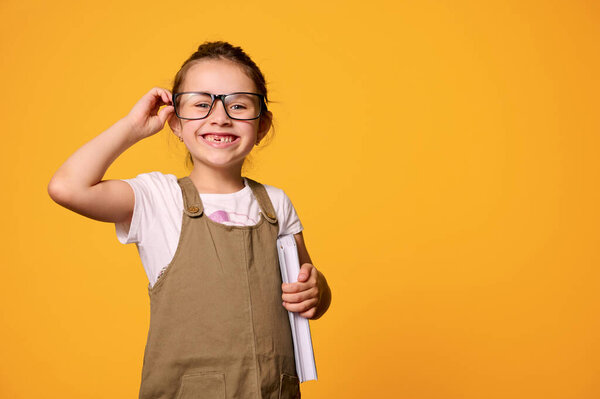 Caucasian mischievous adorable smiling child girl, first grader looking at camera through her stylish eyeglasses, holding a book, isolated over orange studio background with free advertising space.