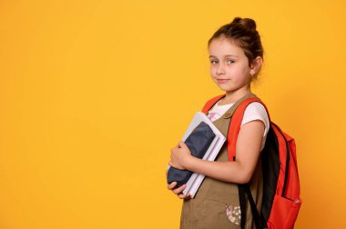 Sırt çantası taşıyan, elinde bir kitap ve kalem çantası olan güzel bir kız kameraya dikkatlice gülümseyen, turuncu stüdyo arka planında izole edilmiş. Akıllı öğrenci. İlkokul çocuğu. Eğitim kavramı