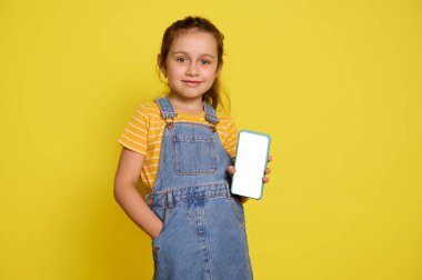 Mavi kot pantolon giyen, elini cebine koyan, kameraya bakıp gülümseyen, beyaz boş dijital dokunmatik ekranlı akıllı cep telefonunu tutan beyaz mutlu bir kız. Mobil uygulama için alanı kopyala