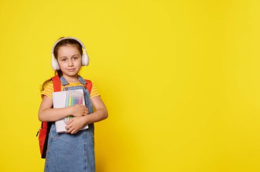 Kablosuz kulaklık takan, sırt çantası taşıyan, kurnazca gülümseyen turuncu stüdyo arka planında izole edilmiş kameraya bakan güzel bir kız çocuğu. Yeni okul yılına hazırlanıyorum. Çocuklar işte. Eğitim