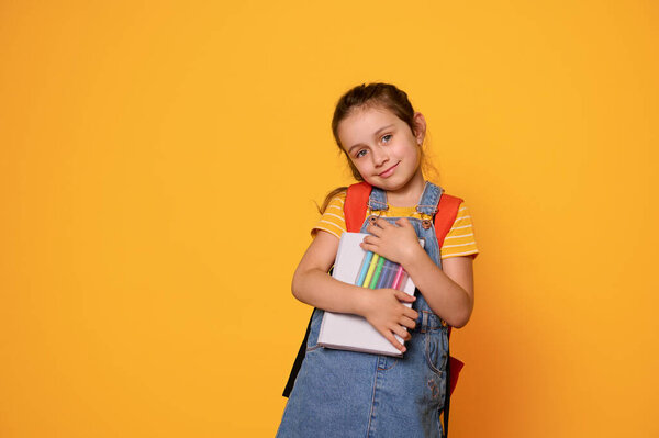 Красивая маленькая девочка с войлочными ручками, с рюкзаком, мило улыбается, смотрит в камеру, изолированная на фоне оранжевой студии. Готовлюсь к новому учебному году. Дети и концепция образования