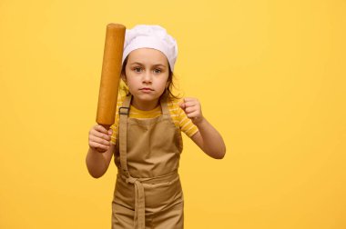 Ahşap bir oklava tutan, yumruklarını sıkan, kameraya bakan, öfkesini dışa vuran, stüdyo sarısı arka planda izole bir şekilde duran, şef üniforması giymiş ciddi bir kız çocuğunun portresi.